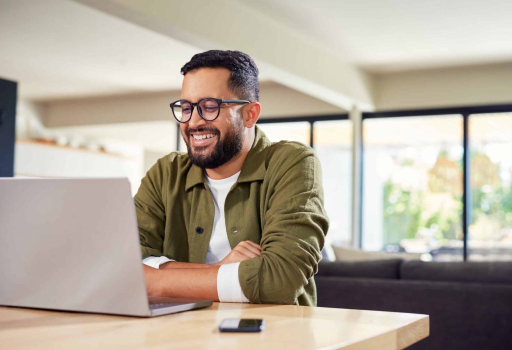Smiling young Indian man sitting at home while using laptop. Multiethnic business man attending meeting online at home while using laptop. Happy mixed race man wearing eyeglasses while using laptop during work from home with copy space.