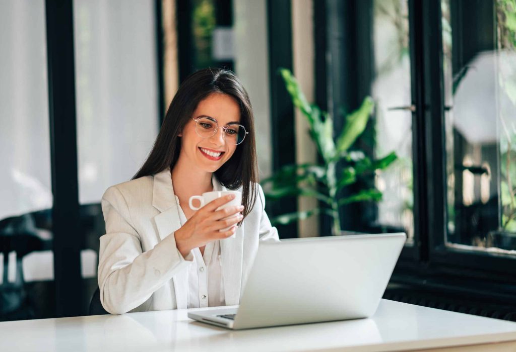 Elegant young woman using laptop and drinking coffee in modern office.