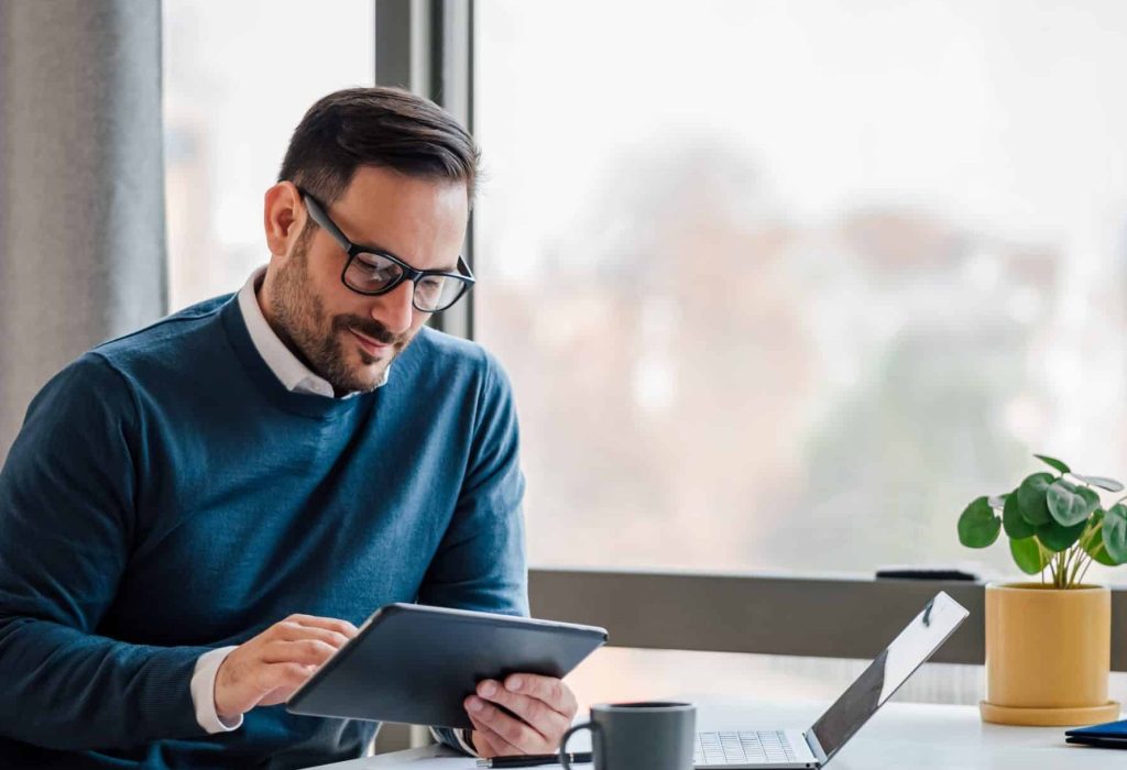 Young businessman using digital tablet while working on laptop in business office. Male professional with wireless computer at desk. He is sitting by window in office.
