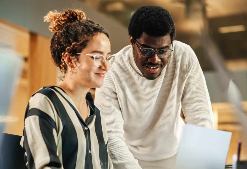 Portrait of Two Creative Colleagues Using Laptop to Discuss Work Project at Office. Young Black Technical Support Specialist Helping Female Customer Relationship Coordinator. Teamwork Concept