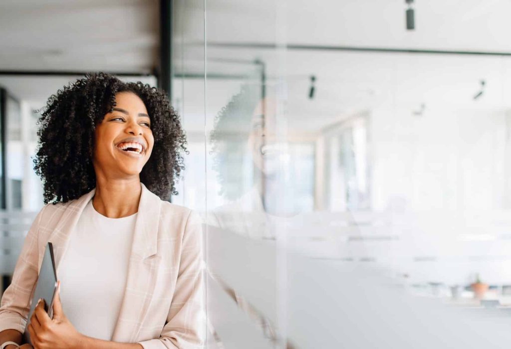 Brazilian businesswoman with an infectious smile standing in a modern office corridor and holding her laptop. Cheerful female employee satisfied with her occupation, feels happy and successful